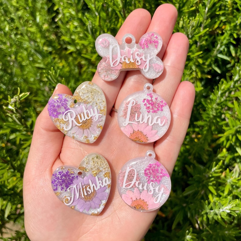 Puede incluir: Una colecci&oacute;n de etiquetas personalizadas para mascotas en forma de coraz&oacute;n y hueso. Cada etiqueta es transparente con flores incrustadas y el nombre de la mascota en cursiva. Los colores incluyen rosa, morado y dorado.