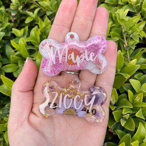 Puede incluir: Dos etiquetas para mascotas en forma de hueso con dise&ntilde;os florales. Una etiqueta tiene flores rosas y el nombre "Maple" en blanco. La otra etiqueta tiene flores moradas y el nombre "Ziggy" en blanco. Ambas etiquetas son transparentes con detalles dorados.