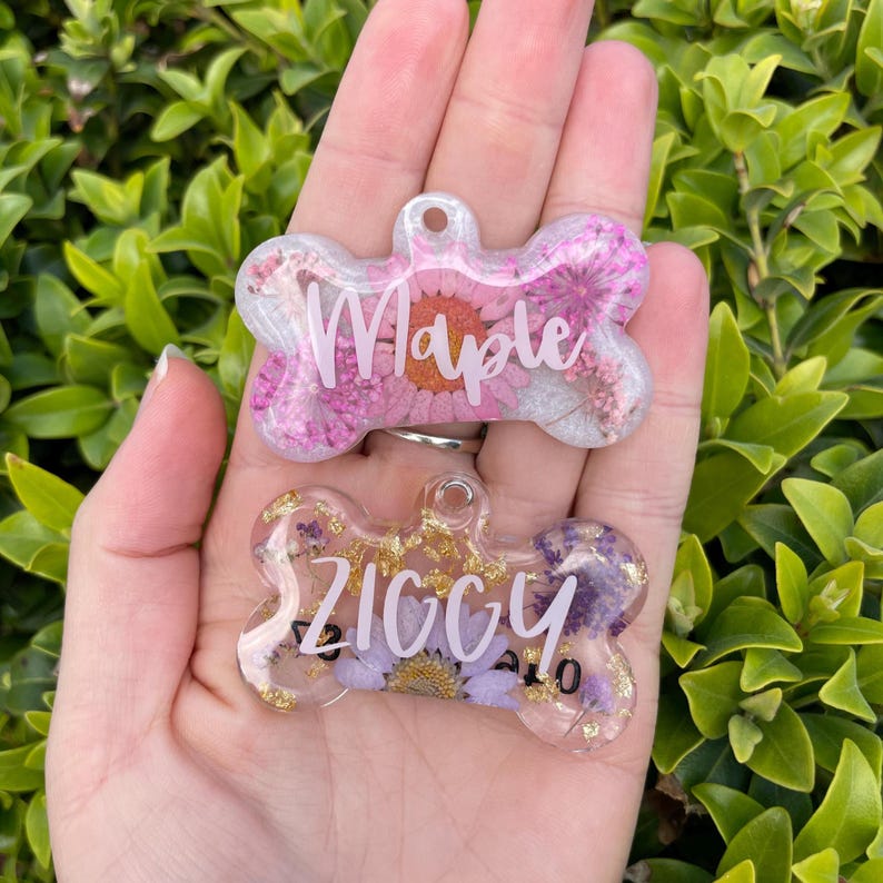 Puede incluir: Dos etiquetas para mascotas en forma de hueso con dise&ntilde;os florales. Una etiqueta tiene flores rosas y el nombre "Maple" en blanco. La otra etiqueta tiene flores moradas y el nombre "Ziggy" en blanco. Ambas etiquetas son transparentes con detalles dorados.