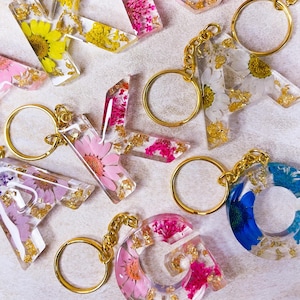 May include: Clear resin letter keychains featuring embedded dried flowers in yellow, pink, white, and blue, accented with gold leaf flakes. Each letter includes a gold-toned metal split ring attached by a short chain for securing keys or accessories.