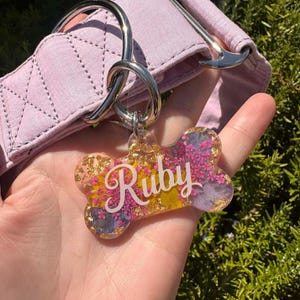 Puede incluir: Una placa para perro en forma de hueso, con el nombre "Ruby" en escritura blanca. La placa est&aacute; llena de flores coloridas y copos de oro, sujeta a un collar para perro lavanda.