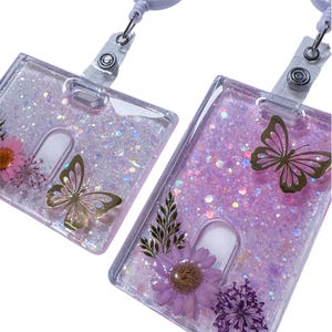 May include: Two rectangular clear resin badge holders with retractable clips. The holders are filled with iridescent glitter and embedded with pressed flowers and gold butterfly silhouettes. One holder features a pink flower, while the other displays a purple flower and a fern leaf.