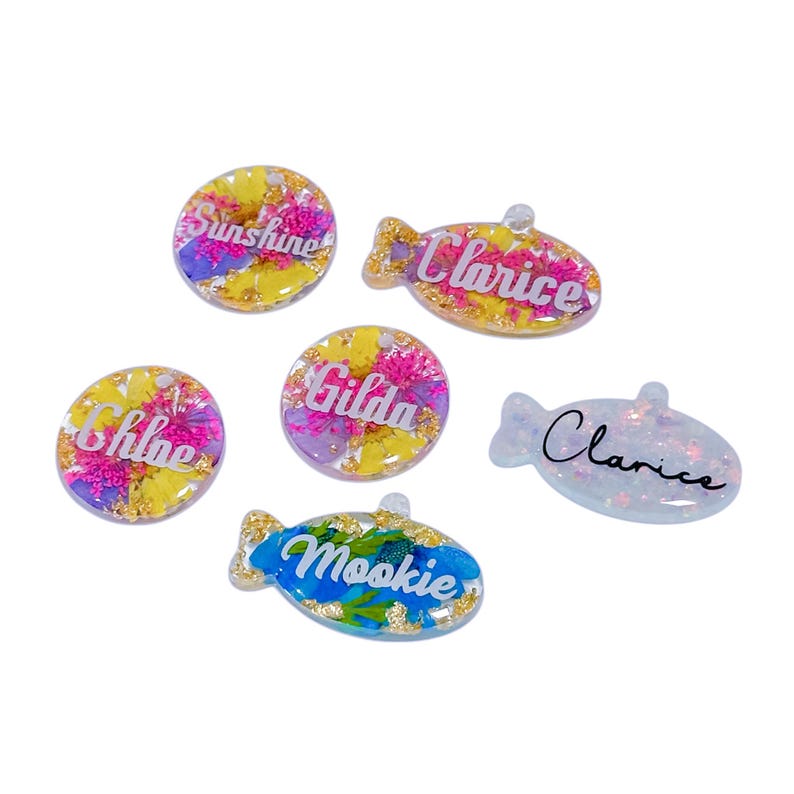 Puede incluir: Un conjunto de colgantes de resina con inclusiones florales y nombres personalizados. Los colgantes tienen varias formas, incluyendo c&iacute;rculos y peces, con nombres como "Sunshine", "Chloe" y "Clarice" en escritura cursiva. La paleta de colores incluye amarillo, rosa y azul.