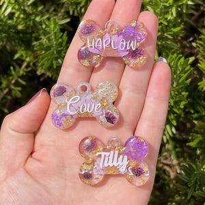 Puede incluir: Tres etiquetas para mascotas en forma de hueso con los nombres Harlow, Cove y Tilly. Cada etiqueta es transparente con flores moradas incrustadas, copos de oro y letras blancas. Las etiquetas tienen un peque&ntilde;o agujero para la fijaci&oacute;n.