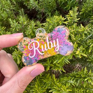 Puede incluir: Una etiqueta transparente para mascotas con forma de hueso con el nombre "Ruby" en escritura blanca. La etiqueta est&aacute; llena de flores secas coloridas y detalles de pan de oro, creando un accesorio &uacute;nico y personalizado.