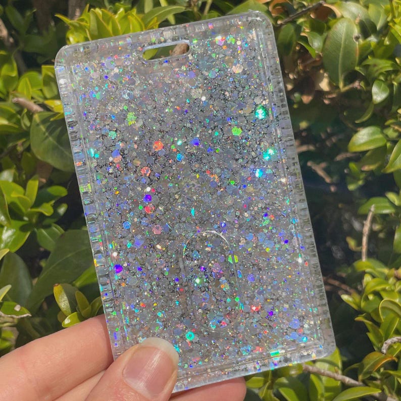 May include: A rectangular clear plastic ID badge holder filled with dense silver and holographic glitter. The holder features a top slot for a lanyard attachment and a thumb slide cutout at the bottom center to assist with card removal.