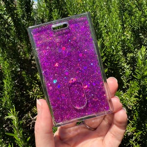 May include: A rectangular clear plastic ID badge holder filled with vibrant purple liquid and a dense mixture of iridescent purple, pink, and blue glitter particles. The holder features a top slot for a lanyard and a central oval thumb slide cutout.