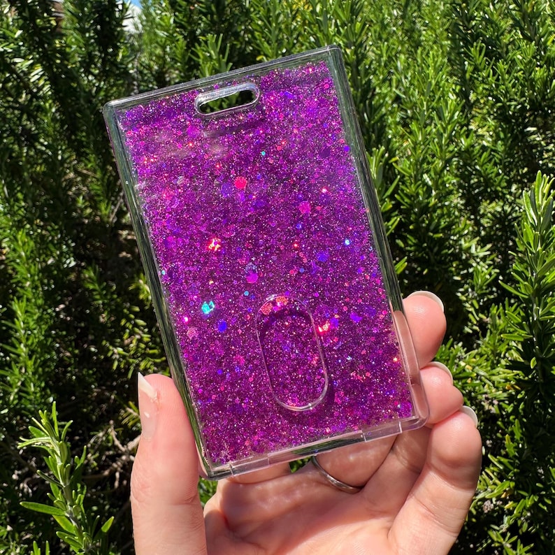 May include: A rectangular clear plastic ID badge holder filled with vibrant purple liquid and a dense mixture of iridescent purple, pink, and blue glitter particles. The holder features a top slot for a lanyard and a central oval thumb slide cutout.