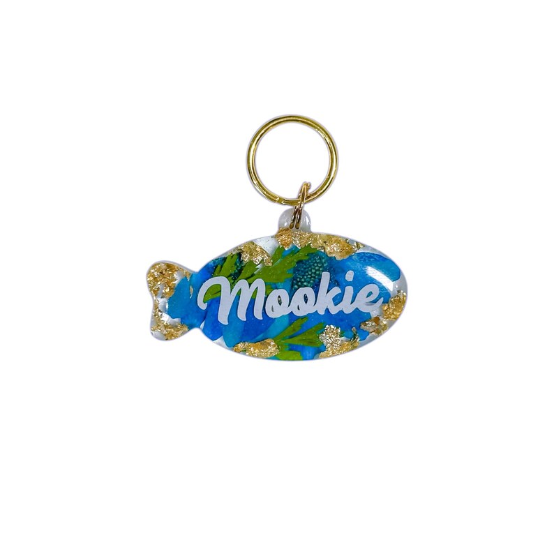 Puede incluir: Una etiqueta para mascotas con forma de pez con el nombre "Mookie" en escritura blanca. La etiqueta presenta dise&ntilde;os florales azules y detalles de pan de oro, sujeta a un anillo dorado.
