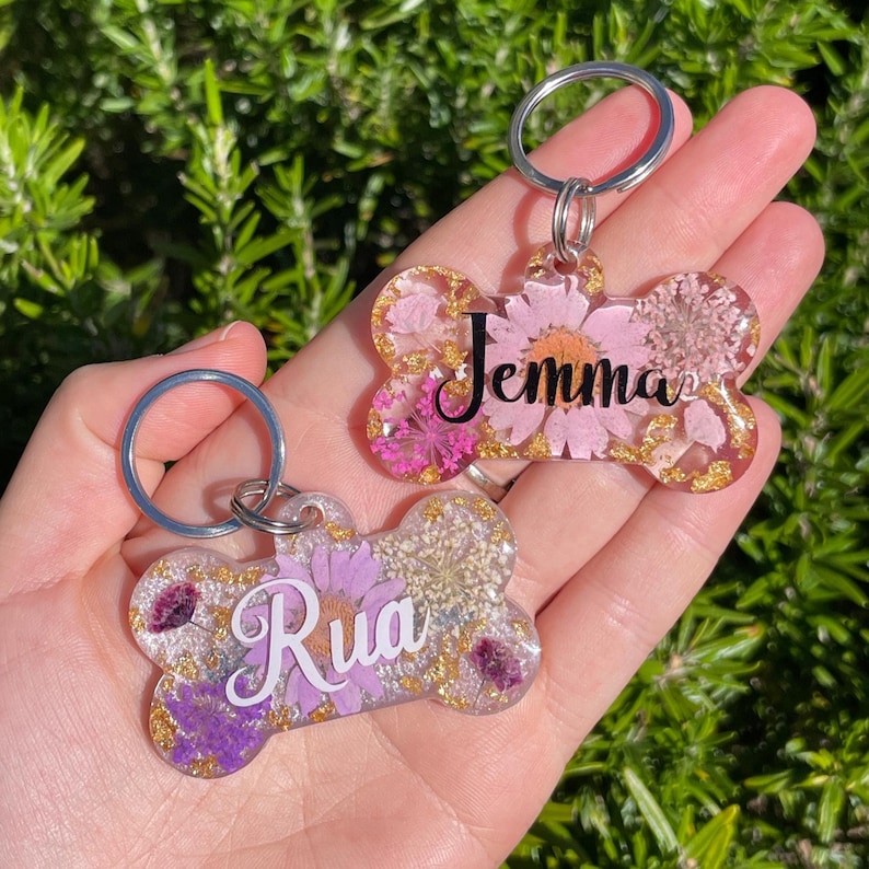 Puede incluir: Dos llaveros en forma de hueso con aros plateados, rellenos de flores secas y hojuelas doradas. Un llavero tiene el nombre "Jemma" en escritura negra, el otro "Rua". Los llaveros est&aacute;n sobre un fondo verde y frondoso.