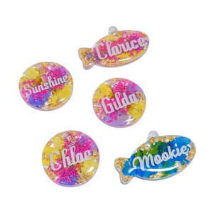 Puede incluir: Cinco colgantes de resina con flores secas y copos de oro. Los colgantes tienen varias formas, incluyendo c&iacute;rculos y peces. Cada colgante tiene un nombre en escritura blanca: "Sunshine", "Clarice", "Gilda", "Chloe" y "Mookie".
