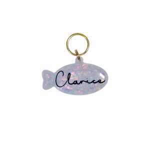 Puede incluir: Una etiqueta para mascotas en forma de pez azul claro con el nombre "Clarice" en escritura negra. La etiqueta tiene un anillo dorado para sujetar. La etiqueta est&aacute; llena de purpurina iridiscente.
