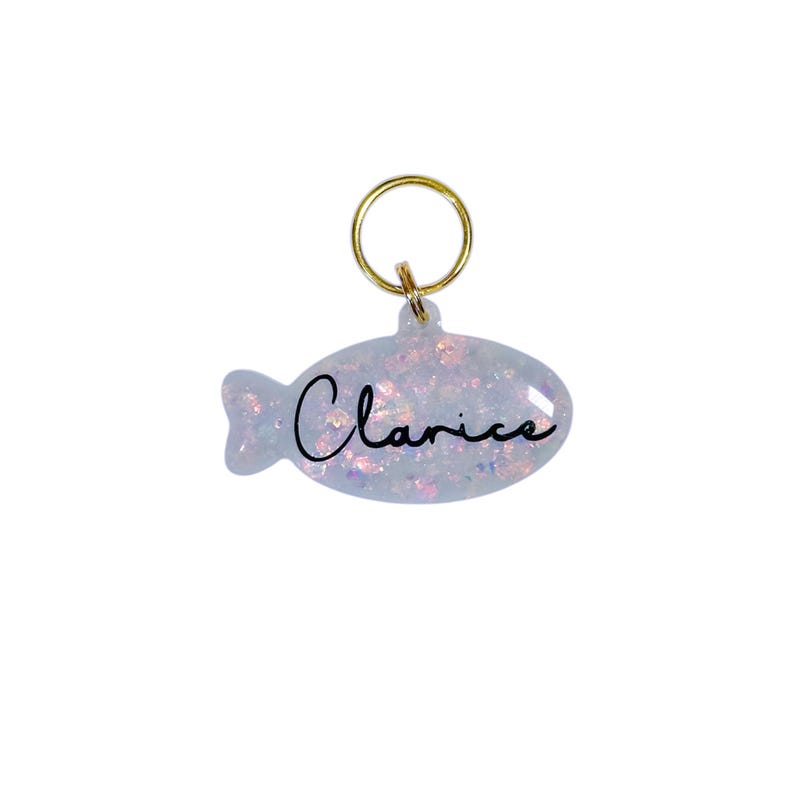 Puede incluir: Una etiqueta para mascotas en forma de pez azul claro con el nombre "Clarice" en escritura negra. La etiqueta tiene un anillo dorado para sujetar. La etiqueta est&aacute; llena de purpurina iridiscente.