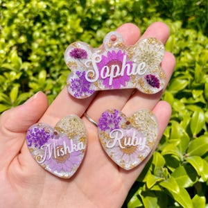 Puede incluir: Tres etiquetas personalizadas para mascotas en forma de hueso y dos corazones. Las etiquetas son de resina transparente con flores secas y copos dorados. Los nombres "Sophie", "Mishka" y "Ruby" est&aacute;n escritos en blanco.
