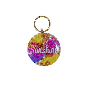 Puede incluir: Un llavero redondo con la palabra "Sunshine" en escritura blanca. El llavero est&aacute; relleno de flores secas amarillas, moradas y rosas, con copos dorados. Un anillo de color dorado est&aacute; unido en la parte superior.