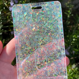 May include: A rectangular clear plastic ID badge holder featuring a dense layer of iridescent, multi-colored glitter embedded within the material. The holder includes a top slot for a lanyard and a central oval cutout for easy card removal.