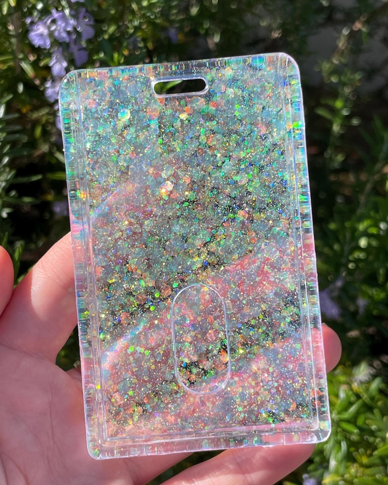 May include: A rectangular clear plastic ID badge holder featuring a dense layer of iridescent, multi-colored glitter embedded within the material. The holder includes a top slot for a lanyard and a central oval cutout for easy card removal.