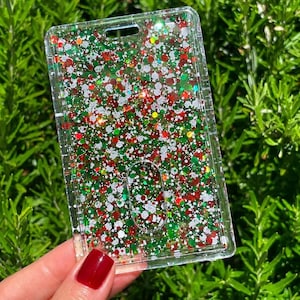 May include: A rectangular clear plastic ID badge holder filled with a festive mix of red, green, and white glitter particles. The holder features a top slot for a lanyard and a central oval cutout, showcasing a vibrant, sparkling holiday-themed design.