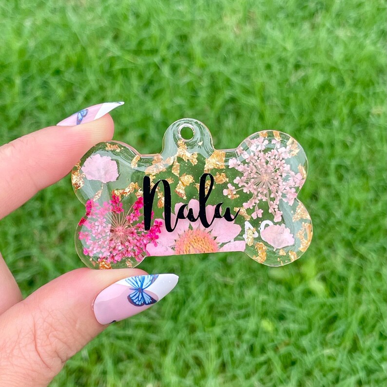 Puede incluir: Una etiqueta transparente para mascotas en forma de hueso con el nombre "Nala" en escritura negra. La etiqueta est&aacute; llena de detalles florales rosas y dorados. La etiqueta se sostiene sobre un fondo de hierba verde.