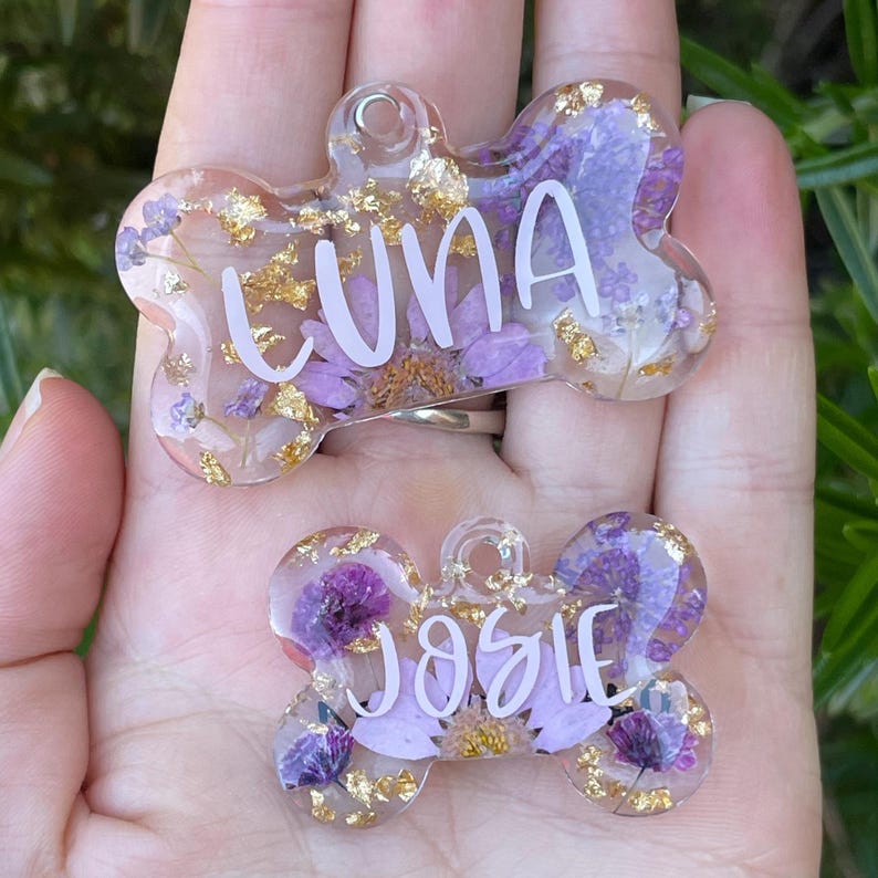 Puede incluir: Dos etiquetas para mascotas en forma de hueso, con resina transparente, que contienen flores moradas y copos dorados. La etiqueta superior dice "LUNA", y la inferior "JOSIE". Ambas tienen un agujero para sujetar.