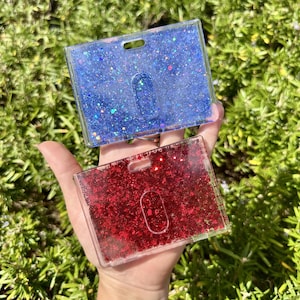 May include: Two rectangular plastic badge holders, one filled with blue glitter and the other with red glitter. Each holder features a clear frame, a top slot for a lanyard, and a central oval cutout for easy card access and identification display.