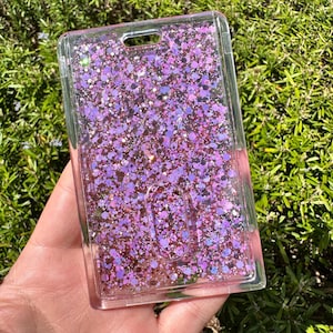 May include: A rectangular clear plastic ID badge holder featuring a liquid-filled interior filled with loose pink and purple glitter particles of various sizes. The holder includes a top slot for a lanyard and a central oval cutout for card access.