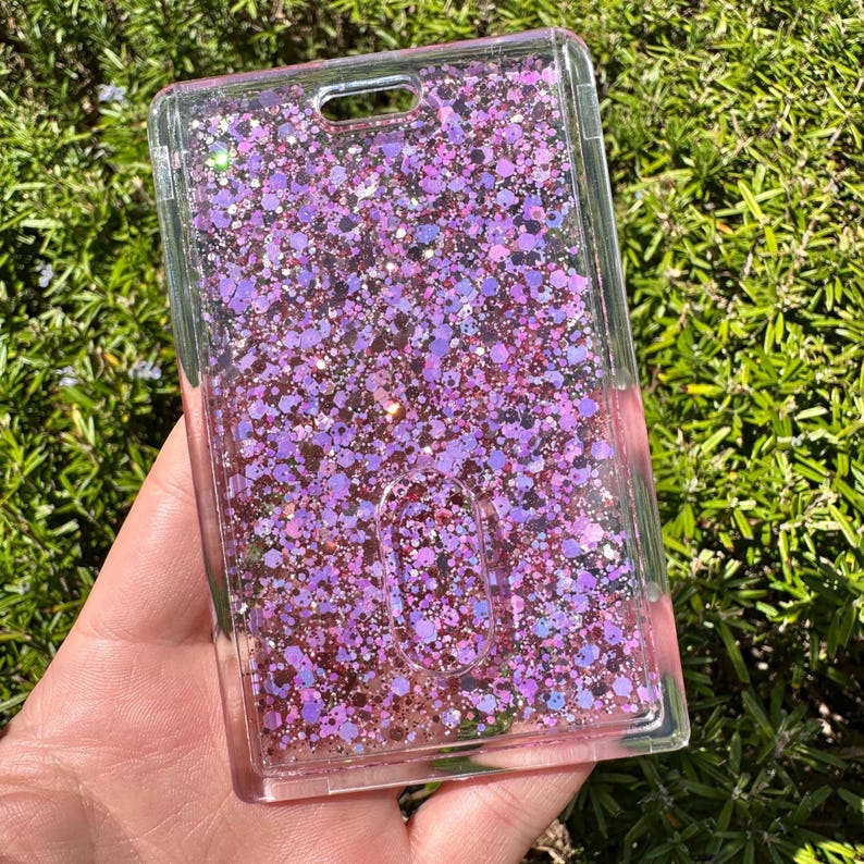 May include: A rectangular clear plastic ID badge holder featuring a liquid-filled interior filled with loose pink and purple glitter particles of various sizes. The holder includes a top slot for a lanyard and a central oval cutout for card access.