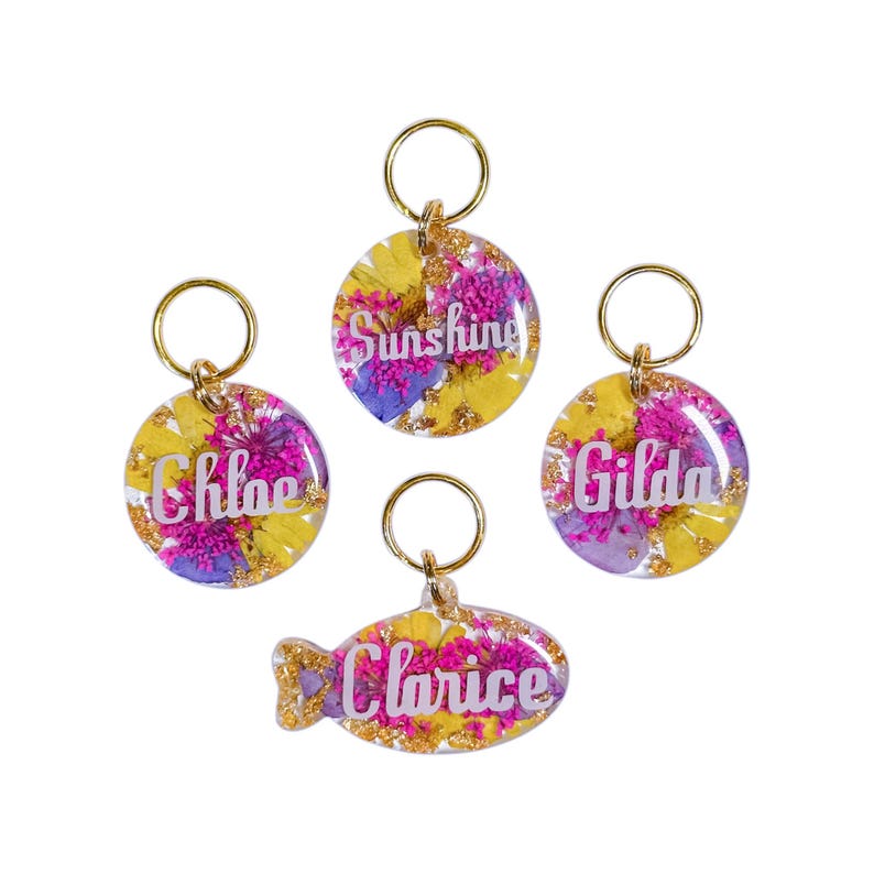 Puede incluir: Cuatro placas de resina para mascotas con anillas de metal dorado. Tres son circulares y una tiene forma de pez. Cada una contiene flores secas amarillas y moradas con copos de oro. Los nombres Chloe, Sunshine, Gilda y Clarice est&aacute;n impresos en blanco.