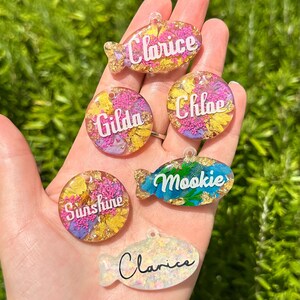 Puede incluir: Una colecci&oacute;n de etiquetas para mascotas coloridas en formas de pez y c&iacute;rculo. Cada etiqueta presenta un nombre en escritura blanca, como "Clarice" y "Mookie". Las etiquetas est&aacute;n llenas de flores secas y copos de oro, sobre un fondo verde.