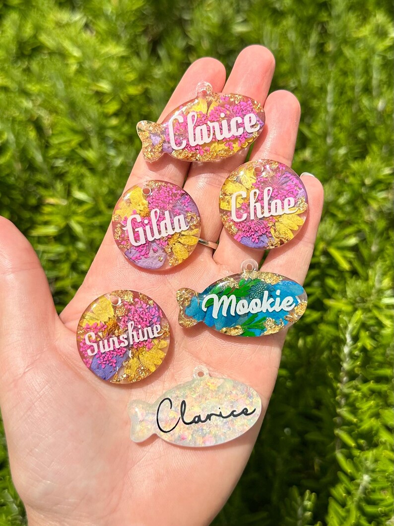 Puede incluir: Una colecci&oacute;n de etiquetas para mascotas coloridas en formas de pez y c&iacute;rculo. Cada etiqueta presenta un nombre en escritura blanca, como "Clarice" y "Mookie". Las etiquetas est&aacute;n llenas de flores secas y copos de oro, sobre un fondo verde.