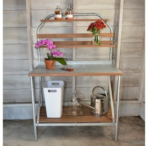 May include: A galvanized metal garden work table with a wooden top and shelves. The table has a white trash can, a glass cloche, and a watering can on the bottom shelf. There are flowers in a glass vase and a potted orchid on the top shelf.