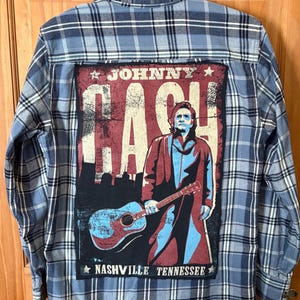 May include: Blue plaid button-down shirt with a graphic of Johnny Cash holding a guitar. The graphic is in red, black, and blue, with the text "JOHNNY CASH" and "NASHVILLE TENNESSEE". The shirt is hanging on a wooden door.