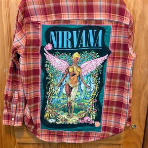 May include: A red and orange plaid flannel shirt with a large patch on the back featuring the band name "NIRVANA" in blue letters. The patch has a colorful illustration of a figure with wings.