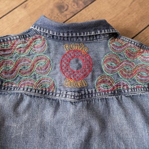 May include: Close-up of a denim jacket's back, featuring colorful embroidered designs. The upper back has the words "Funky Bitch" embroidered in red and yellow, with a red circle design. The jacket has a classic collar and a faded blue color.