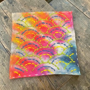 May include: Square textile art piece with a tie-dye effect in shades of pink, orange, yellow, and blue. The design features a repeating pattern of stitched arcs in yellow, blue, and green thread. The edges are finished with white stitching.