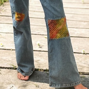 May include: Light blue denim jeans with colorful embroidered patches on the knees. The jeans have a flared leg and are worn barefoot on a wooden surface.