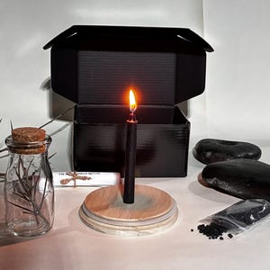 Puede incluir: Una vela negra encendida sobre una base de madera, parte de un conjunto ritual. El conjunto incluye una botella de vidrio con corcho, una caja negra, piedras negras y una pequeña bolsa de gránulos negros. También hay un pergamino.