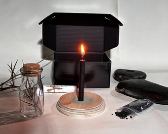 May include: A black candle with a lit flame sits on a wooden base, part of a ritual set. The set includes a glass bottle with a cork, a black box, black stones, and a small bag of black granules. A scroll is also present.