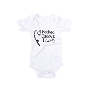 May include: White baby onesie with short sleeves and a black graphic of a fishing rod and the text "I hooked Daddy's heart".