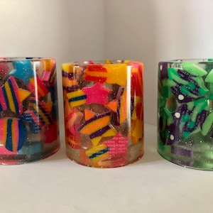 May include: Three clear glass containers filled with colorful, glittery, geometric shapes. The shapes are in shades of pink, yellow, green, blue, and purple.