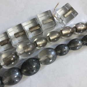 May include: A strand of clear glass beads with silver accents, and a strand of dark gray glass beads. The clear beads are square and round, and the dark gray beads are round.