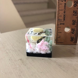 May include: A small, clear cube filled with a miniature underwater scene. The cube contains white sand, a pink coral, a yellow and black seashell, and a red and white string. The cube is sitting on a wooden surface.