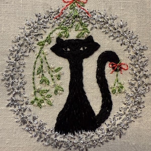 May include: Embroidered artwork featuring a black cat within a silver and green wreath. The cat has a long, slender body and tail, with a sprig of mistletoe tied with a red bow. The wreath is adorned with silver leaves and green mistletoe.