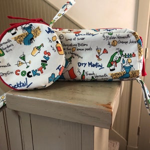 May include: Two white fabric zippered pouches with red trim. The pouches are decorated with colorful illustrations of cocktails and cocktail recipes. The larger pouch has the text "Dry Martini" and "French Vermouth". The smaller pouch has the text "Cocktails" and "Vermouth Gordon's Gin".