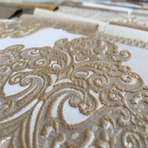 Könnte beinhalten: Nahaufnahme eines weißen Stoffes mit einem erhabenen, komplizierten, goldfarbenen Muster. Das Muster ist ein sich wiederholendes Design aus Spiralen und Kurven.