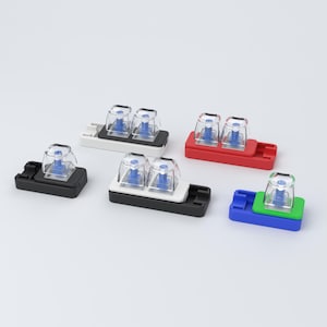 May include: A collection of mechanical keyboard switch testers featuring clear plastic keycaps with visible blue internal stems. The testers are mounted on rectangular bases in black, white, red, blue, and green, arranged in a scattered layout.