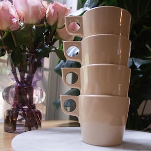 Könnte beinhalten: Ein Stapel von vier beigefarbenen Keramikbechern mit kreisförmigen Griffen. Die Becher stehen auf einem weißen Marmor-Servierbrett. Im Hintergrund steht eine Vase mit rosa Rosen, neben einem Fenster.