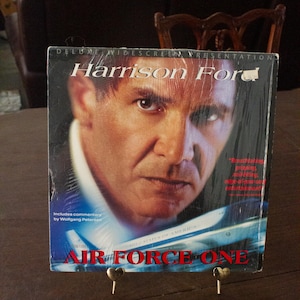 May include: A VHS tape case for the movie Air Force One, featuring Harrison Ford on the cover. The case is blue and has a red title with the text "DELUXE WIDESCREEN PRESENTATION" and "AIR FORCE ONE".