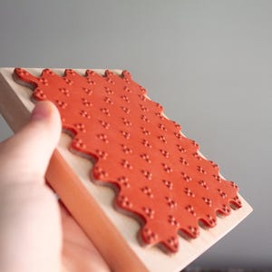May include: A red rubber stamp with a repeating pattern of small dots is mounted on a rectangular wooden block. The stamp has a wavy, scalloped edge. The stamp is held at an angle.