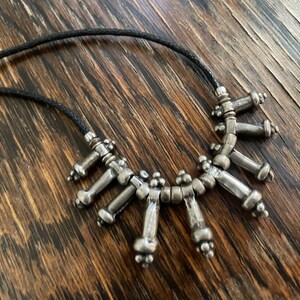 Könnte beinhalten: Eine silberne Halskette mit geometrischem Design. Die Halskette besteht aus kleinen, miteinander verbundenen Silberteilen, die eine Reihe von Punkten bilden. Die Halskette ist auf einer schwarzen Schnur aufgefädelt.